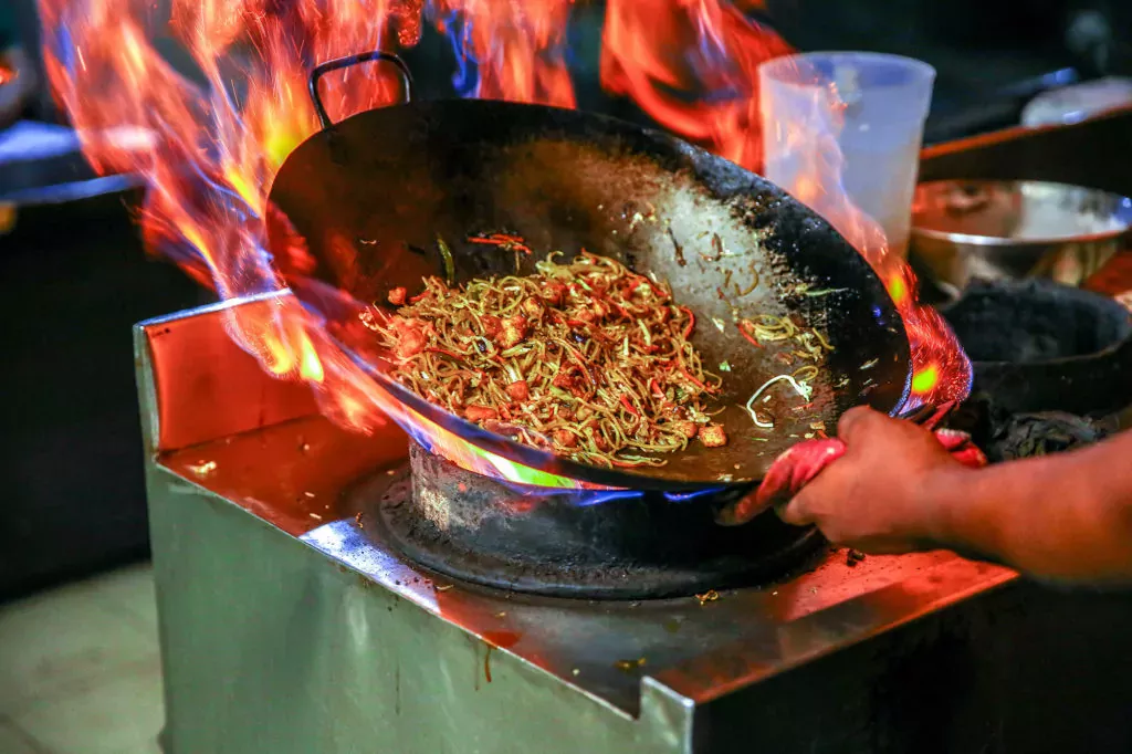 Large Carbon Steel wok with stir fry over huge flames in a commercial kitchen. These guys know there's no comparison between cast iron wok vs carbon steel. It's carbon steel all the way.