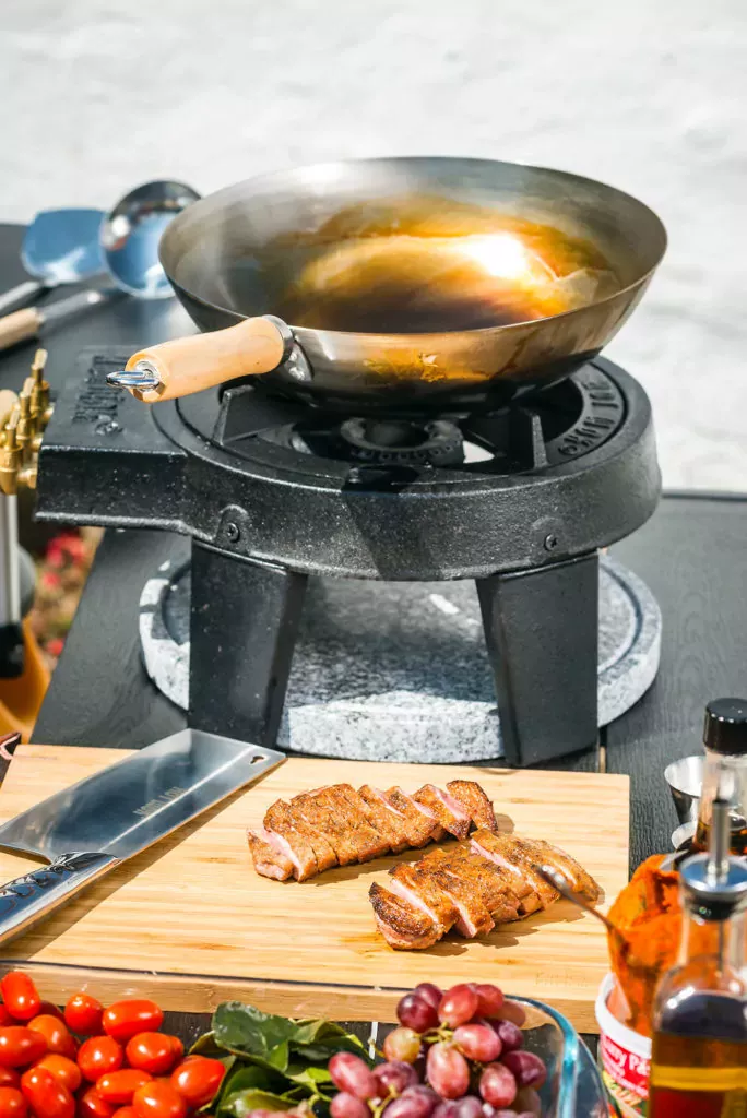 Steel wok on an propane outdoor wok burner, with assorted meat and veggies on a cutting board with a knife