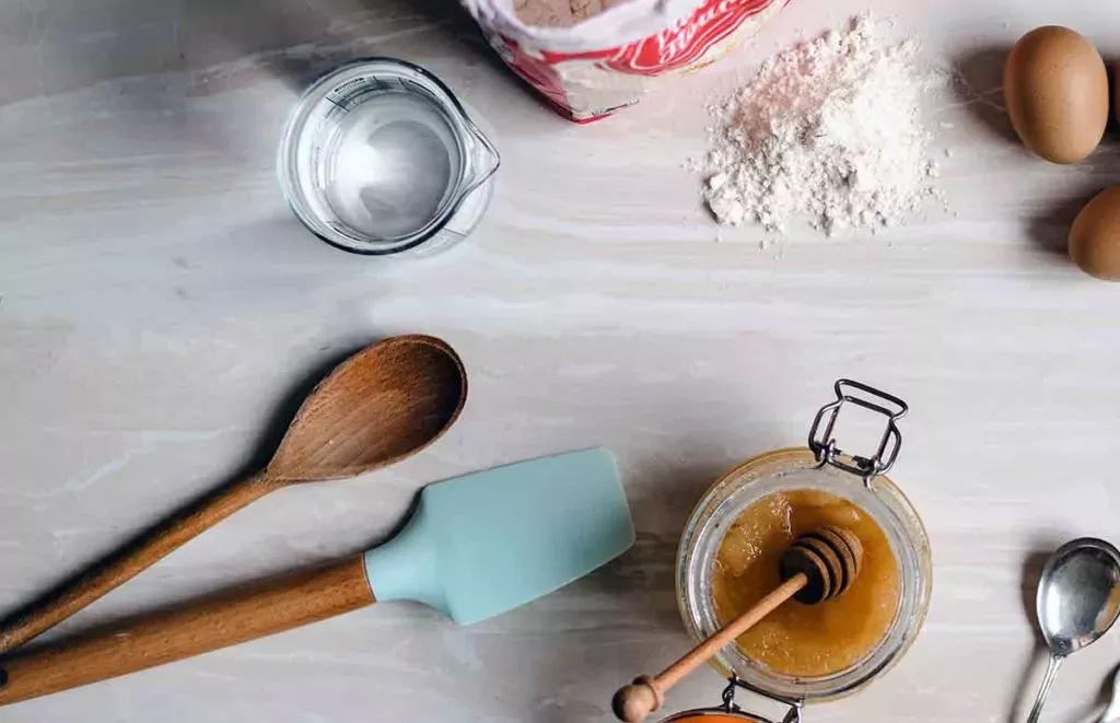 Wooden spoon and rubber scraper next to various ingredients to bake cookies on the counter